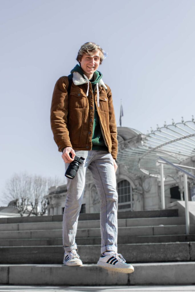 Photo de profil de Romain VIDAL, souriant avec un appareil photo à la main. Photo de Romain Vidal, souriant avec un appareil photo à la main et descendant les escaliers. Vidéaste & Photographe à Clermont-Ferrand.