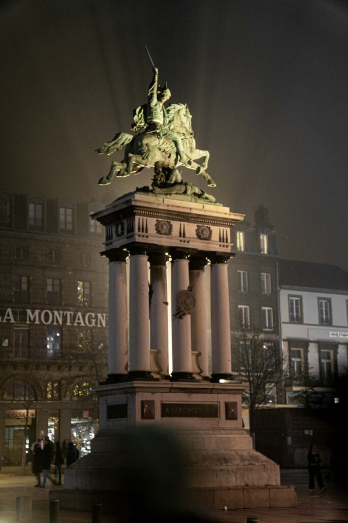 Statue de Vercingétorix à Clermont-Ferrand, éclairée pendant la nuit. Photo de Romain Vidal, souriant avec un appareil photo à la main et descendant les escaliers. Vidéaste & Photographe à Clermont-Ferrand.